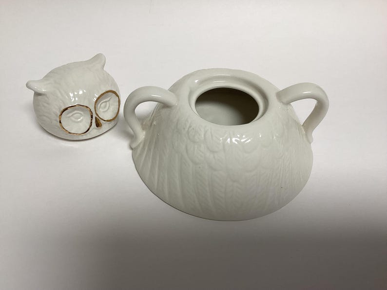 Puede incluir: Un azucarero de cer&aacute;mica blanca con forma de b&uacute;ho con tapa a juego. La tapa es una peque&ntilde;a cabeza de b&uacute;ho con detalles dorados en los ojos. El cuenco tiene dos asas y un dise&ntilde;o de plumas texturizadas.