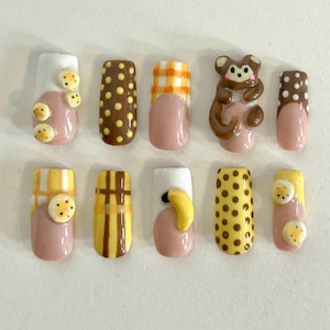 Könnte beinhalten: Ein Set aus zehn künstlichen Nägeln mit verschiedenen Designs. Die Nägel weisen eine Mischung aus Farben auf, darunter Rosa, Weiß, Braun und Gelb. Einige Nägel haben Polka Dots, Karomuster oder 3D-Affen- und Bananen-Designs. Die Nägel sind unterschiedlich lang.