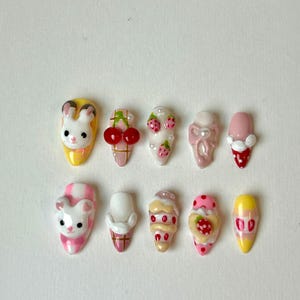 May include: A collection of ten artificial nails with various designs. The designs include a bunny, cherries, strawberries, and ice cream cones. The nails are painted in a variety of colors, including pink, yellow, white, and red. The nails are oval-shaped.