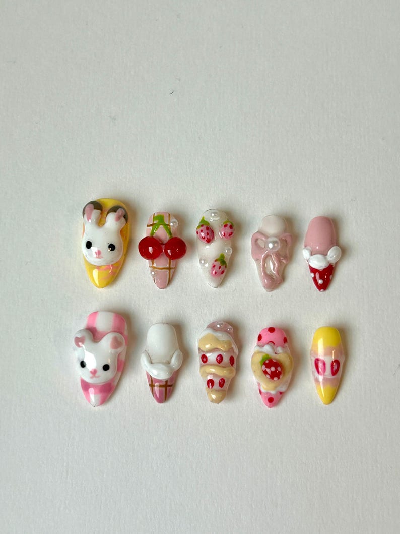 May include: A collection of ten artificial nails with various designs. The designs include a bunny, cherries, strawberries, and ice cream cones. The nails are painted in a variety of colors, including pink, yellow, white, and red. The nails are oval-shaped.