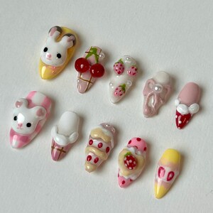 May include: A collection of ten artificial nails with various designs. The designs include a bunny, a bear, cherries, strawberries, and ice cream cones. The nails are painted in a variety of colors, including pink, yellow, white, and red.