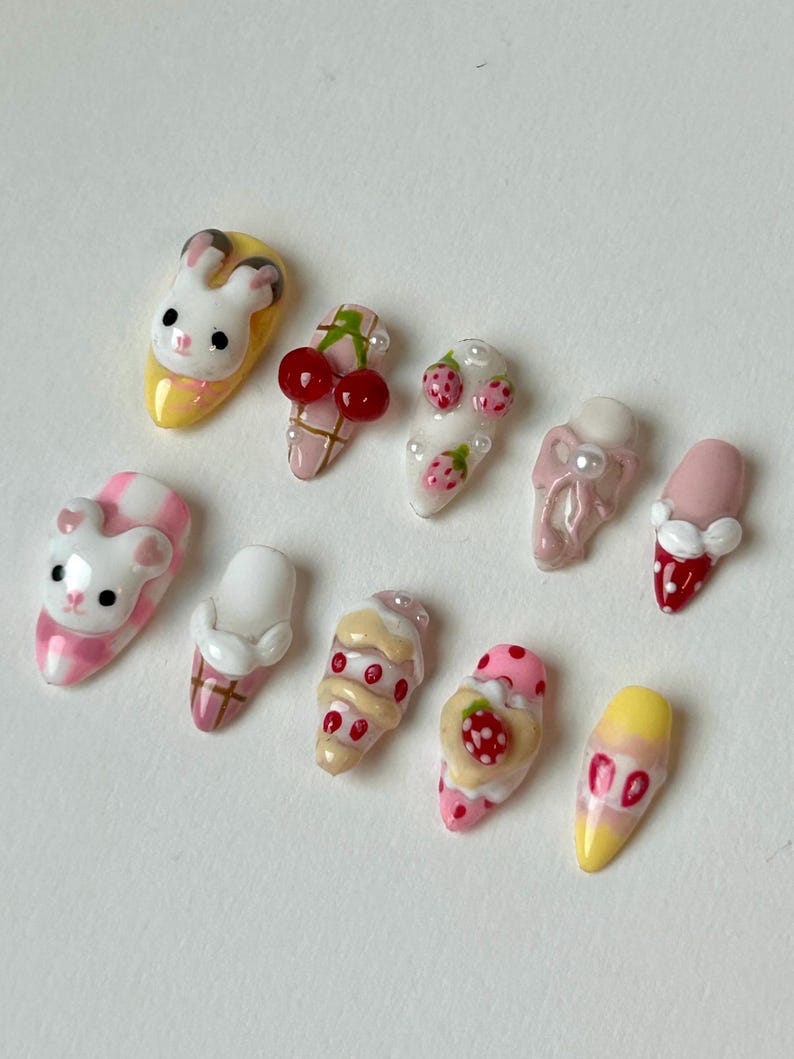 May include: A collection of ten artificial nails with various designs. The designs include a bunny, a bear, cherries, strawberries, and ice cream cones. The nails are painted in a variety of colors, including pink, yellow, white, and red.