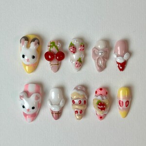 May include: A collection of ten press-on nails with various designs. The designs include a bunny, cherries, strawberries, and ice cream cones. The nails are painted in pastel colors, including pink, yellow, and white. Some nails have pearl accents.