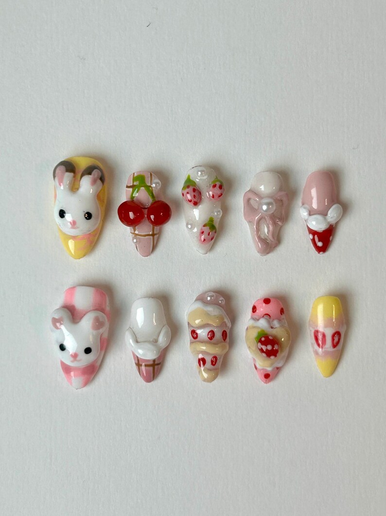 May include: A collection of ten press-on nails with various designs. The designs include a bunny, cherries, strawberries, and ice cream cones. The nails are painted in pastel colors, including pink, yellow, and white. Some nails have pearl accents.