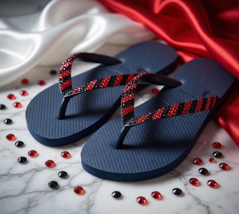 Peut inclure: Tongs bleu marine avec des lani&egrave;res noires orn&eacute;es de strass rouges et noirs. Les sandales sont pr&eacute;sent&eacute;es sur une surface en marbre avec des pierres pr&eacute;cieuses rouges et noires &eacute;parpill&eacute;es, et un tissu satin&eacute; rouge et blanc en arri&egrave;re-plan.
