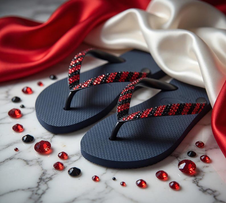 Peut inclure: Une paire de tongs bleu marine avec des lani&egrave;res orn&eacute;es de strass rouges et noirs altern&eacute;s. Des pierres pr&eacute;cieuses rouges et noires sont dispers&eacute;es autour des sandales, sur un fond de tissu de satin rouge et blanc.