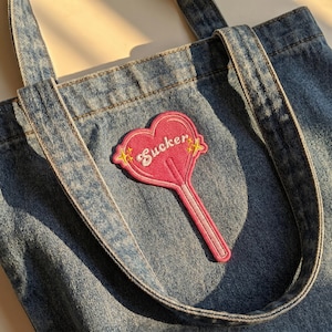May include: A denim tote bag with a pink heart-shaped lollipop patch that reads "Sucker" in cursive. The patch has yellow star accents. The bag has two denim handles and is made of a light blue denim material.
