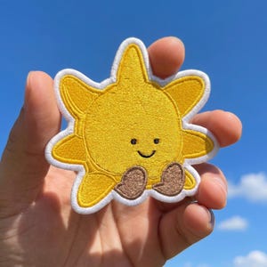 May include: A yellow embroidered sun patch with a white border, featuring a simple black stitched face with two small eyes and a curved smile, and two brown rounded feet positioned at the bottom of the circular sun shape with radiating rays.