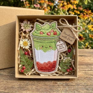 May include: An embroidered patch shaped like a cup of matcha with strawberry hearts, featuring a smiling face, inside a cardboard gift box filled with shredded paper, dried flowers, and a tag that reads Handmade with Love and Strawberry Hearts Matcha.