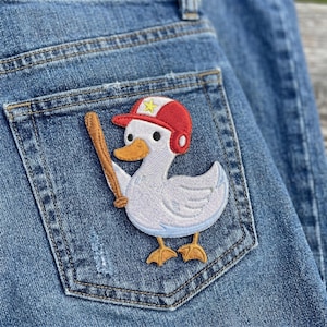 May include: A denim blue jean pocket featuring an embroidered patch of a white duck wearing a red baseball cap with a yellow star and holding a wooden baseball bat. The duck has an orange beak and feet.