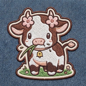 May include: An embroidered patch featuring a brown and white cow with pink flowers on its head, a small yellow bell collar, and green grass in its mouth. The cow stands on a patch of green grass with tiny flowers, all outlined in dark brown thread.