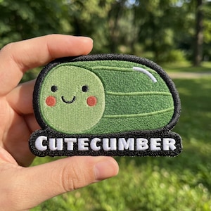 May include: An embroidered patch featuring a green cucumber character with a smiling face, pink cheeks, and black eyes. The patch has a black border and the white text CUTECUMBER printed on a black base, creating a distinct graphic design.