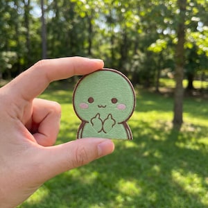 May include: A green embroidered patch featuring a cartoon character with a round head and body. The character has brown eyes, pink cheeks, and is giving the middle finger with both hands. The patch has a brown border.