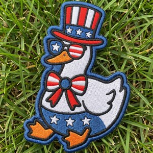 May include: Embroidered patch featuring a patriotic goose wearing a top hat, sunglasses, and a bow tie, all in red, white, and blue. The goose has orange feet and a beak, and is set against a green grass background. The patch is outlined in blue.
