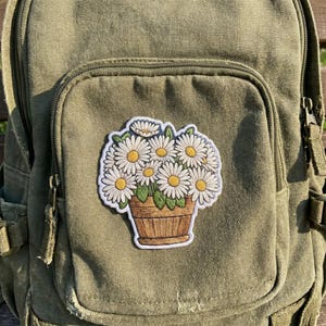May include: An olive green canvas backpack featuring a stitched fabric patch of a bouquet of white daisies with yellow centers arranged in a brown wooden bucket. The patch has a white border and is centered on the front pocket of the backpack.