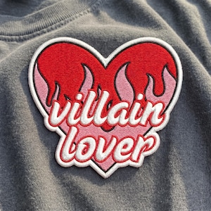 May include: Embroidered patch in the shape of a heart with red flames and the words "villain lover" in a cursive font. The patch has a white border and is set against a gray fabric background.