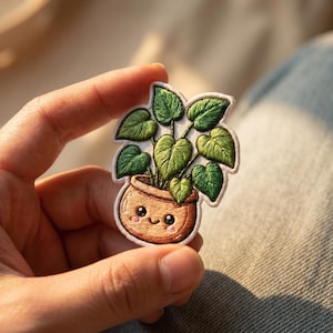 May include: Embroidered patch featuring a potted plant with large green leaves. The plant is in a brown pot with a smiling face. The patch has a white border and is held in a hand.