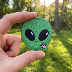 May include: A circular embroidered patch featuring a green alien face with large black oval eyes, two small black dots for nostrils, and a small pink tongue sticking out from the side of the mouth, all outlined with a thick green border.