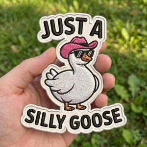 May include: Embroidered patch featuring a white goose wearing a pink cowboy hat and sunglasses. The patch has the words "JUST A" above the goose and "SILLY GOOSE" below it, all in black lettering with a white border.