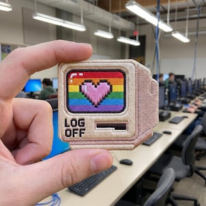 May include: A beige embroidered patch shaped like a vintage computer monitor. The screen displays a pixelated rainbow background with a pink heart in the center. Below the screen, the text LOG OFF is stitched in black, next to a black slot.