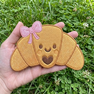 May include: Embroidered patch in the shape of a croissant with a smiling face and a pink bow. The patch is a golden-yellow color with brown details and a heart-shaped mouth.