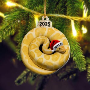 May include: A Christmas ornament featuring a coiled yellow snake with white spots. The snake wears a Santa hat with the name "Bella". The ornament has a gold string and the year "2025" at the top. The background is a blurred Christmas tree.