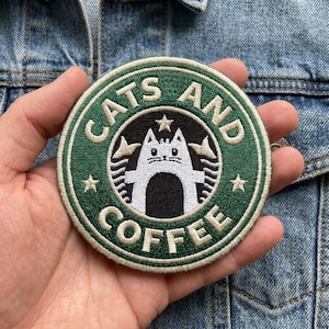 May include: A round, green and white embroidered patch with the words "CATS AND COFFEE" in a circular design. The patch features a white cat silhouette in the center, resembling a coffee shop logo. The patch is held in a hand.