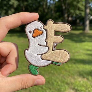 May include: Embroidered patch featuring a cartoon duck embracing a tan letter. The duck has a white body, orange beak, and green feet. The patch is held up against a green background.