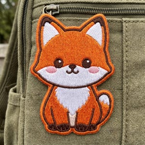 May include: Embroidered patch featuring a cartoon fox with orange fur, white chest, and brown accents. The fox has large, dark eyes and pink cheeks. The patch is attached to a green canvas backpack, showcasing its potential use as an accessory.