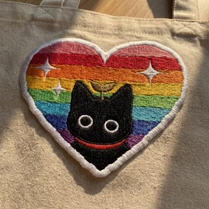 May include: A beige tote bag featuring a heart-shaped patch with a rainbow design. The patch has a black cat face with a red smile and a small green sprout. Two white starbursts are on the patch.