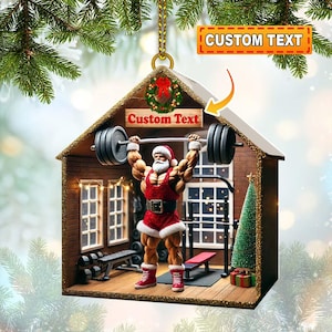 May include: A Christmas ornament shaped like a gym, featuring a muscular Santa Claus lifting weights. The ornament has a red and white Santa suit, a small Christmas tree, and the text "Custom Text".