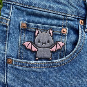 May include: An embroidered patch featuring a gray bat with light pink inner wings and ears, positioned on the front pocket of blue denim jeans. The bat has black eyes, a small nose, and two tiny white fangs visible on its face.