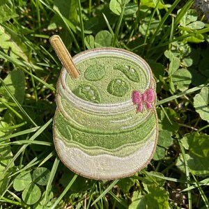 May include: An embroidered patch shaped like a glass jar filled with a green liquid, featuring a tan straw, several green circular elements, and a small pink bow attached to the side, with a white base and a tan stitched border.