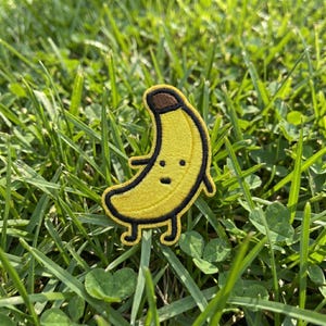 May include: A yellow banana patch with a smiling face and black outline. The patch has a brown stem and small arms and legs. The patch is set against a background of green grass.