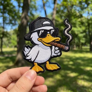 May include: Embroidered patch featuring a cartoon duck wearing sunglasses and a backward baseball cap, holding a cigar. The duck has white feathers, a yellow beak and feet, and is set against a blurred green background.