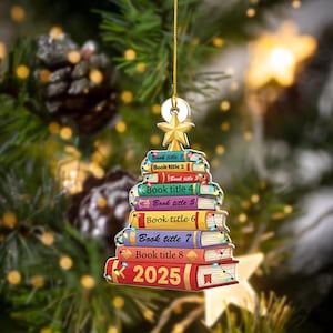 May include: A Christmas tree ornament shaped like a stack of books. Each book is a different color and has the text "Book title" and a number. The bottom book has "2025" printed on it. The ornament is hanging from a gold string and has a gold star at the top.