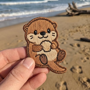 May include: Embroidered patch featuring a cartoon otter holding a seashell. The otter has brown fur, black eyes, and a tan belly. The patch is outlined in brown thread. The background shows a sandy beach and ocean.