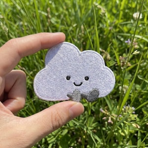 May include: A light gray cloud-shaped embroidered patch with a smiling face and small gray arms. The patch has a white border and is held against a background of green grass.
