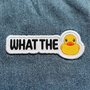 May include: Embroidered patch with the phrase "WHAT THE" in black capital letters on a white background. A yellow cartoon duck with an orange beak is on the right side of the patch. The patch is on a blue denim background.