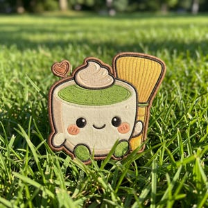 May include: Embroidered patch featuring a cute matcha latte with a smiling face, whipped cream, and a heart. A yellow bamboo whisk is next to the cup. The patch is on a green grassy background.