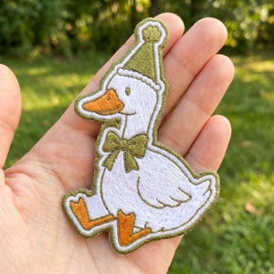 May include: Embroidered patch featuring a cartoon goose wearing a green party hat and bow tie. The goose has an orange beak and feet, with white feathers and a green outline. The patch is held in a hand against a blurred green background.