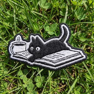 May include: Embroidered patch featuring a black cat lounging on a book with a cup of coffee. The cat has white eyes and is outlined in white. The book and coffee cup are also white with black details. The patch is on a green grassy background.