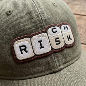 May include: Olive green baseball cap with the word "RISK" embroidered on the front in a keyboard-style design. The letters are white with a brown border. The cap has a curved brim and a fabric strap.