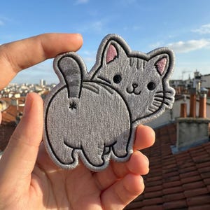 May include: Embroidered gray cat patch with pink inner ears and black eyes. The cat is depicted from the rear, with its tail raised. The patch is designed with a playful and humorous aesthetic, suitable for clothing or accessories.
