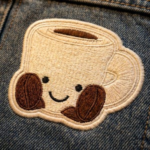 May include: Embroidered patch featuring a smiling coffee cup with two coffee beans. The patch is beige with brown accents and black eyes and a curved mouth. The patch is sewn onto a blue denim fabric.