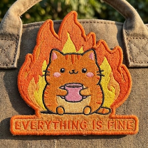May include: Embroidered patch featuring a cartoon orange cat holding a pink mug, surrounded by orange and yellow flames. The patch reads "EVERYTHING IS FINE" in orange text on a rectangular banner. The patch is attached to a tan canvas bag.