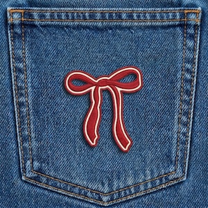 May include: Close-up of a blue denim pocket with a red and white bow patch. The bow is a classic design with a white outline, adding contrast against the denim's texture and color. The pocket has visible stitching.