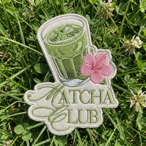 May include: An embroidered patch featuring a glass of iced green matcha tea with a pink hibiscus flower. The text MATCHA CLUB is stitched in green thread below the glass. The patch has a white border and is shaped to follow the design contours.
