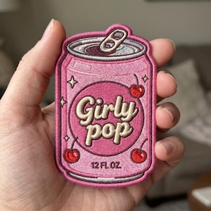 May include: Pink embroidered patch in the shape of a soda can with the words "Girly Pop" in a cursive font. The patch features cherries and stars, and the text "12 FL OZ."