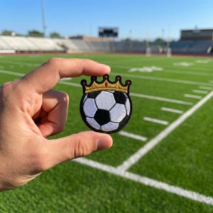 May include: Embroidered patch featuring a soccer ball design with a gold crown. The patch has a black border and is held against a green football field background. The soccer ball has white and black pentagons.